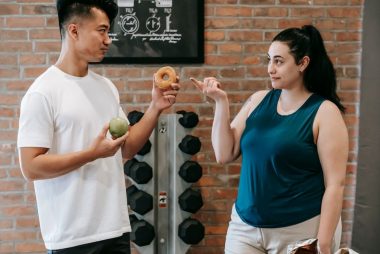 Side view of Asian personal instructor with apple and doughnut offering healthy food to plus size woman while standing in gym